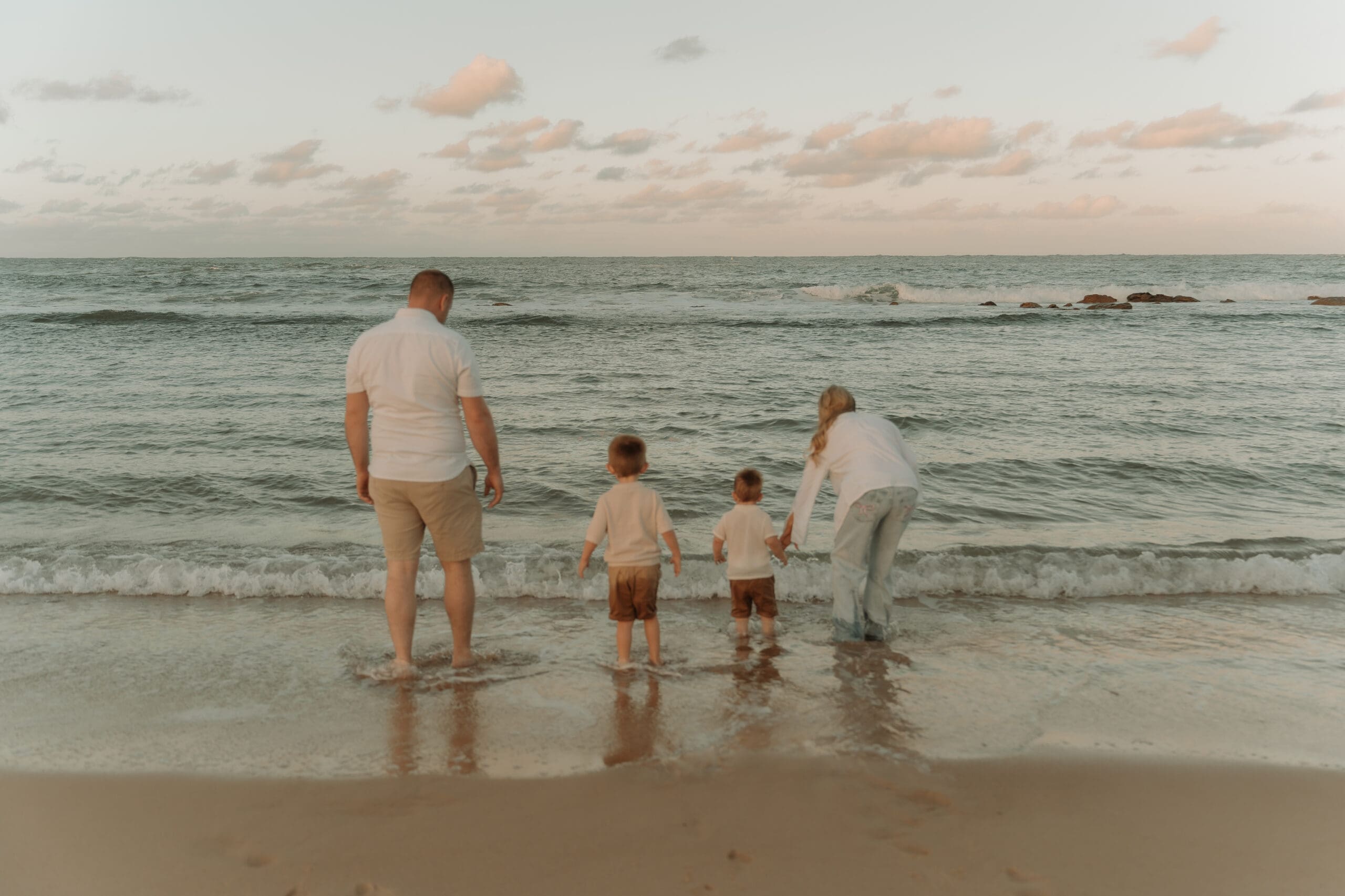 A family of four stand together at the shoreline of the beach, dipping their toes in the water.